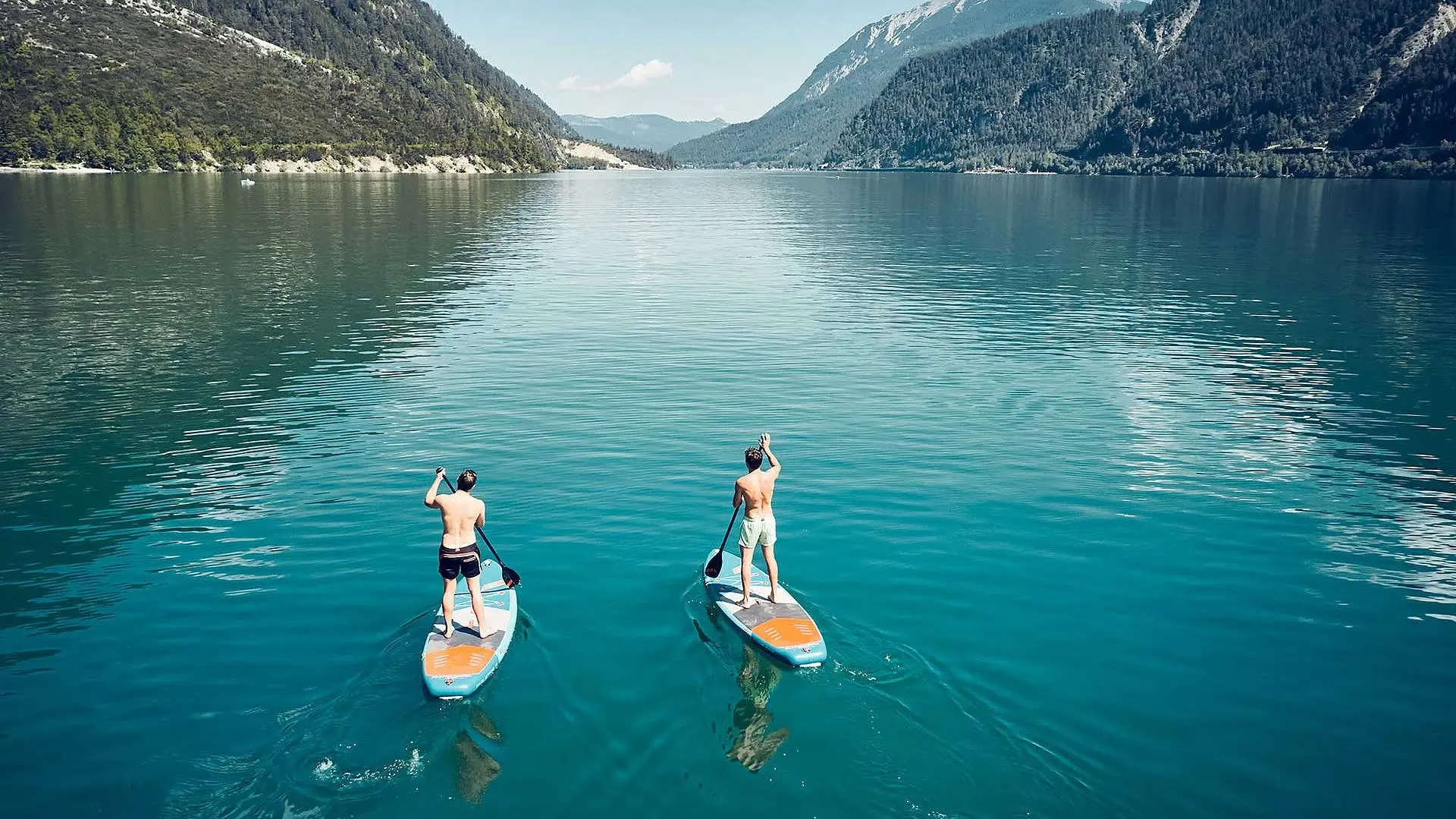 Zwei Männer auf Paddleboards auf einem See mit Bergen im Hintergrund.