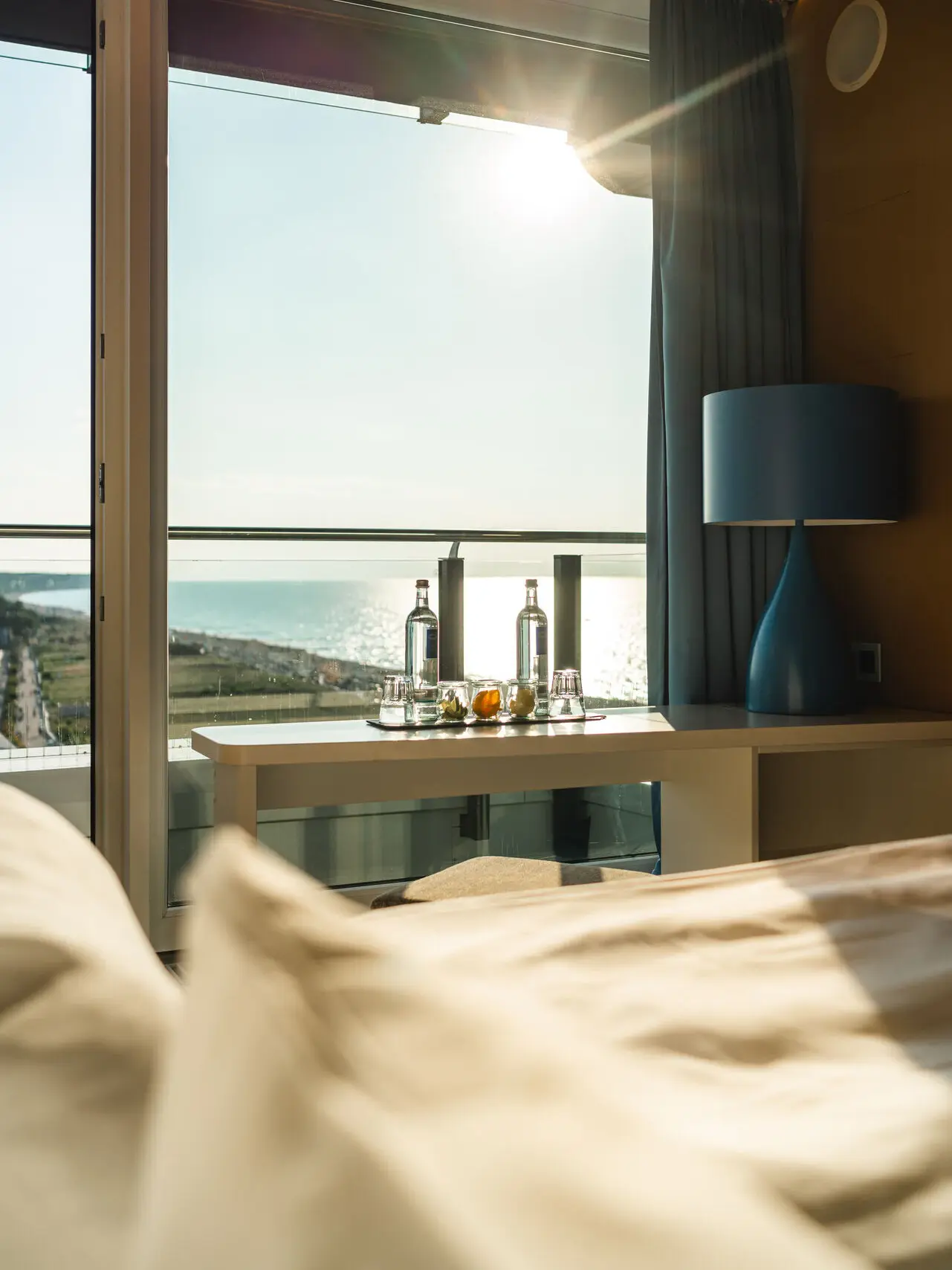 Ein Ausschnitt eines Hotelzimmers mit einem gemütlichen Bett und kleinem Schreibtisch mit Wasserflaschen und Gläsern vor einem großen Fenster, das die Ostsee überschaut.