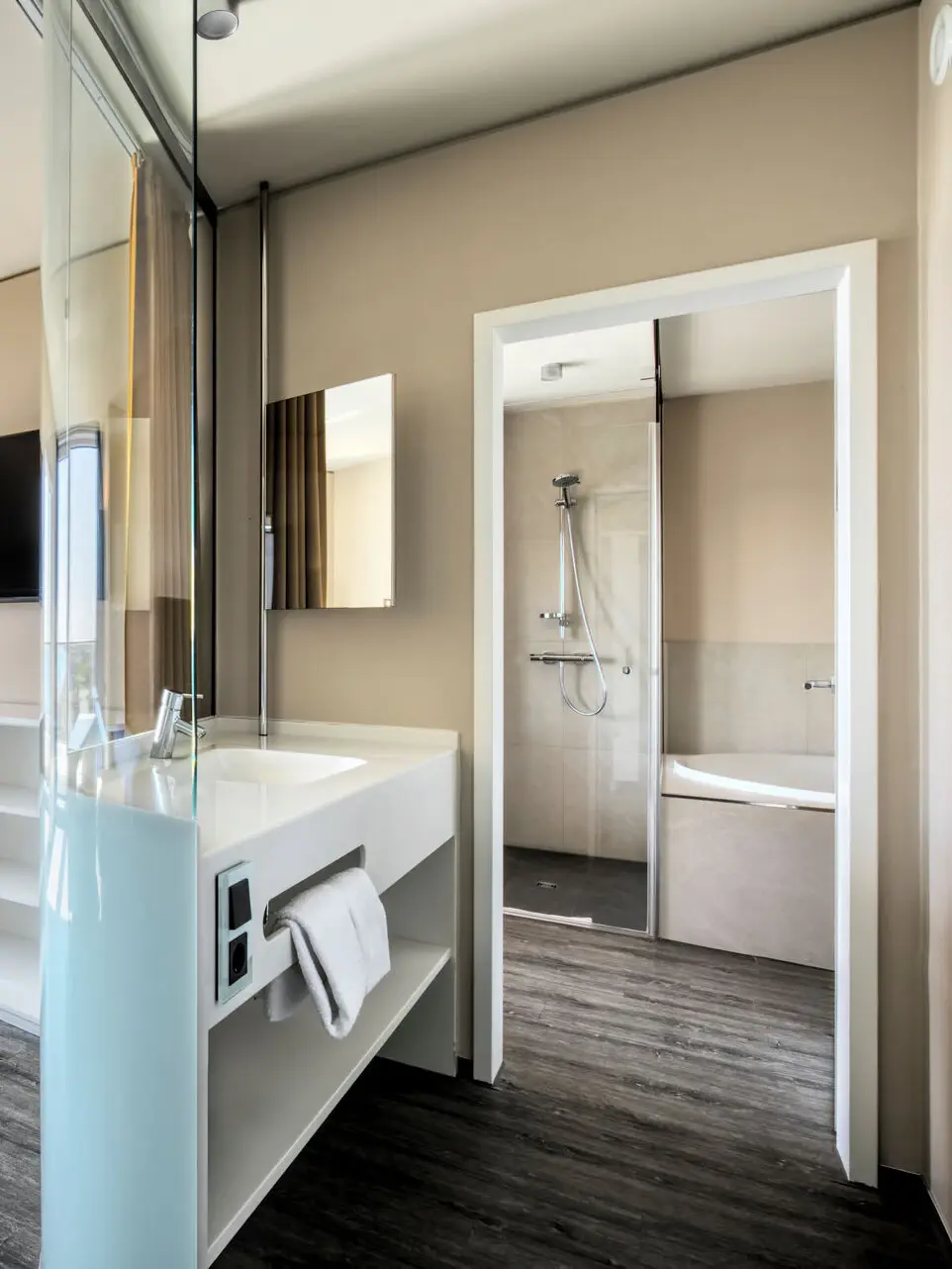 Ein helles Badezimmer mit großem Waschbecken neben einer Glasdusche und dunklem Holzboden.