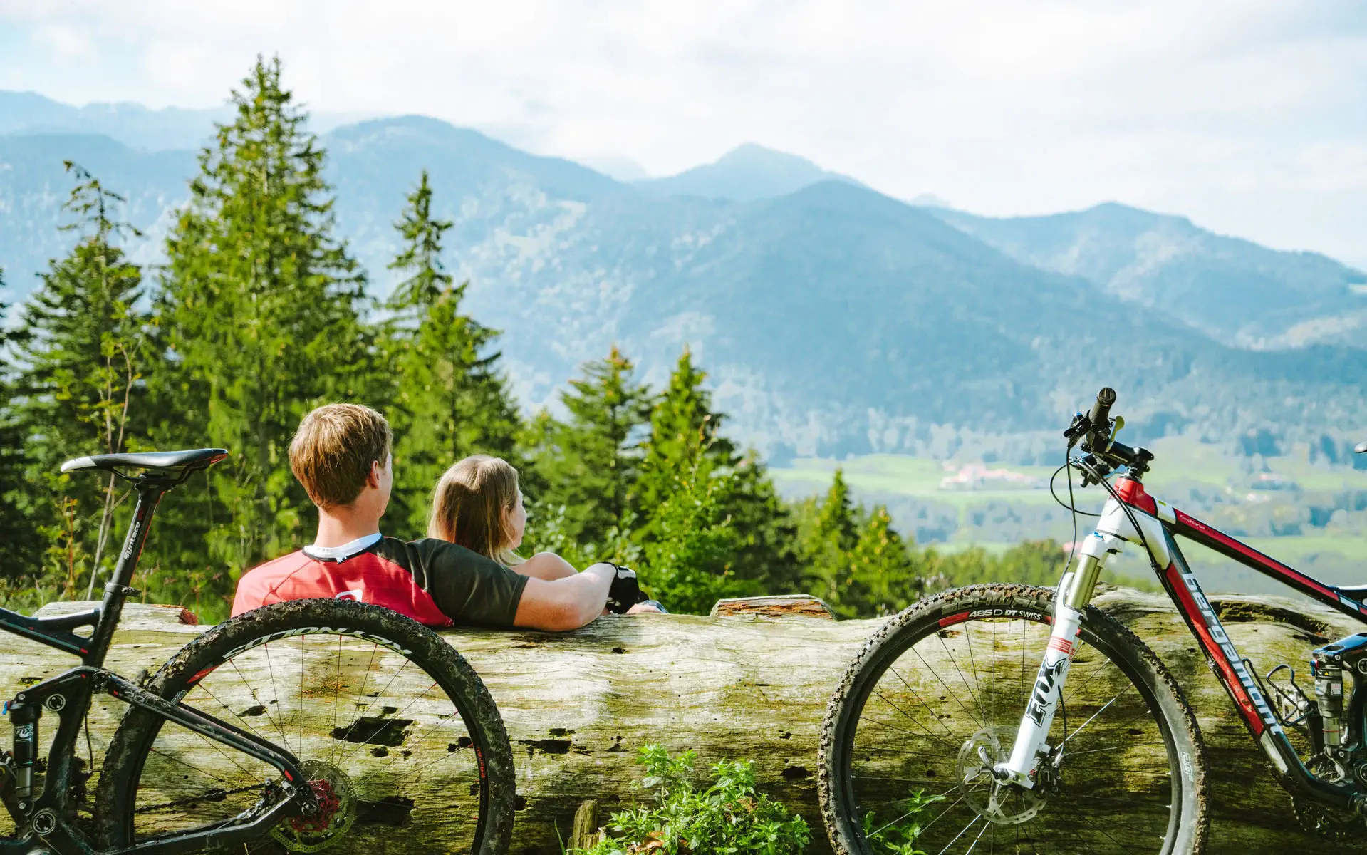 Ein Mann und eine Frau sitzen an einen umgekippten Baumstamm gelehnt auf einer Wiese in den Bergen und schauen in die Ferne. Neben ihnen stehen ihre Fahrräder und die Sonne scheint.
