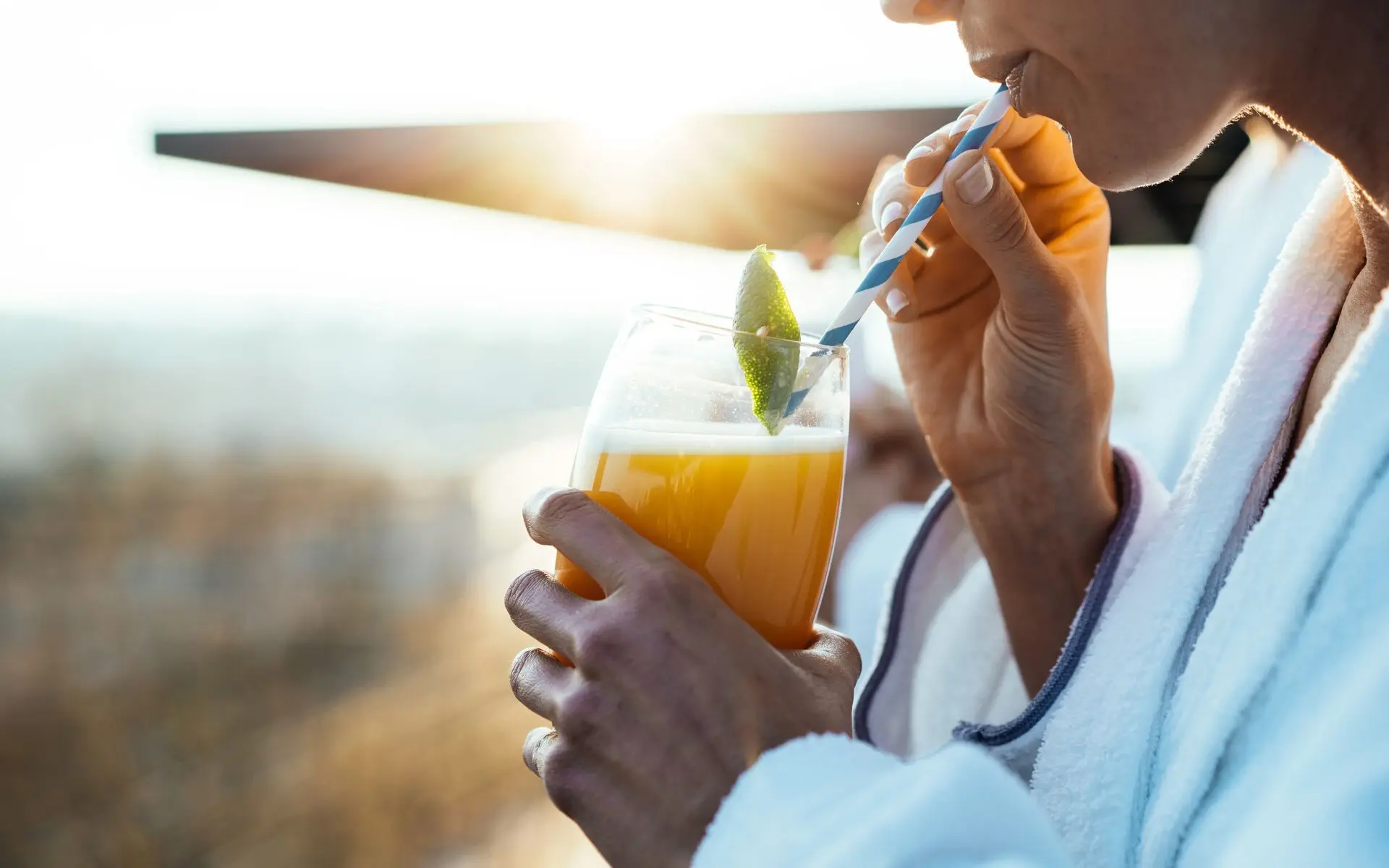 Eine Frau in Bademantel steht auf einer Terrasse in der Morgensonne und trinkt einen orangenen Saft.