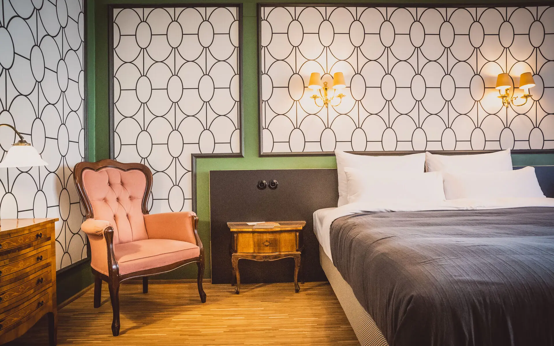 HENRI Wien Charmantes Zimmer im HENRI Wien mit einem großzügigen Doppelbett, stilvoller Tapete im Retro-Design, einem eleganten rosafarbenen Sessel, einem antiken Nachttisch und warmem, gemütlichem Ambiente.