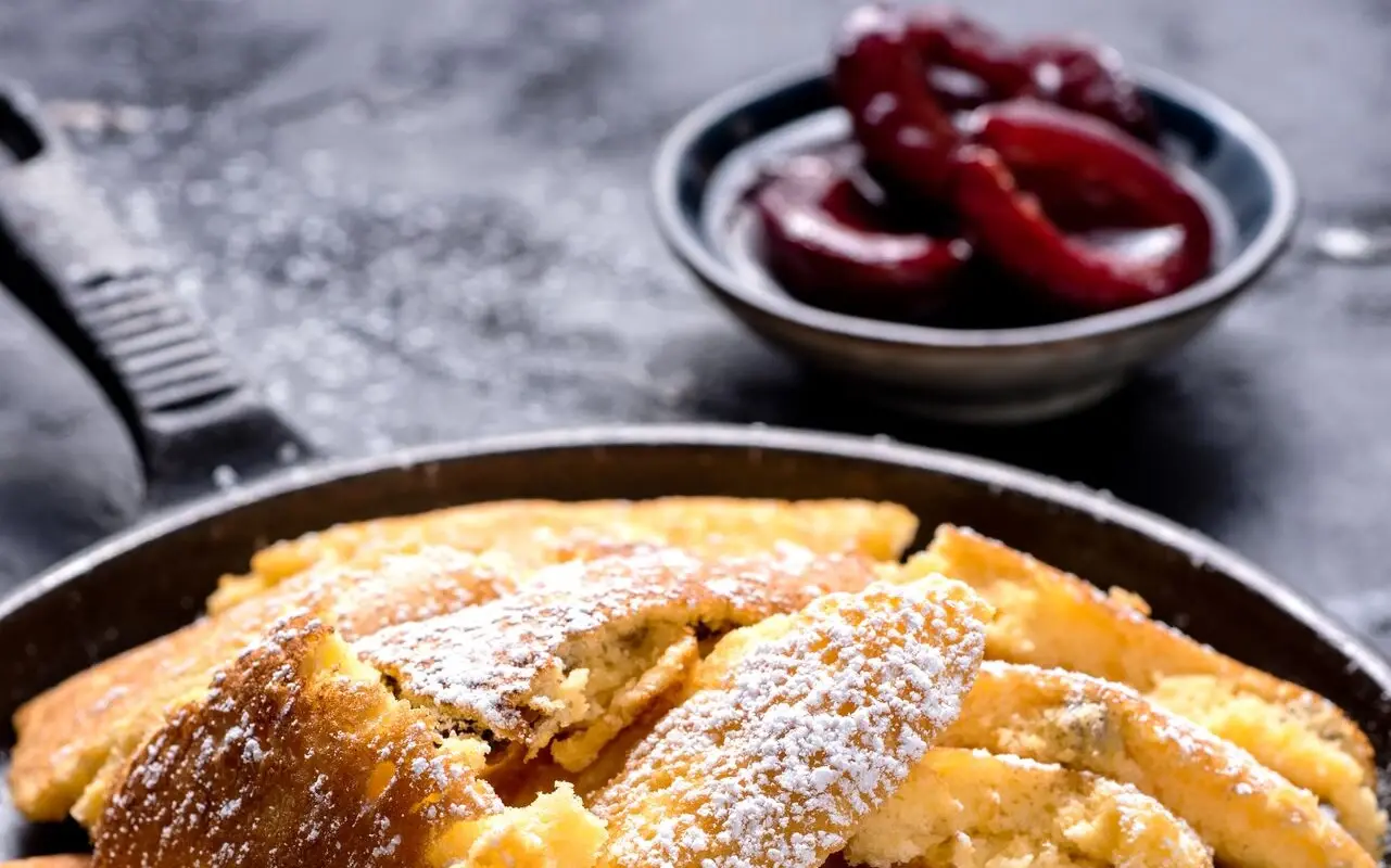 Kaiserschmarrn Eine dunkle Pfanne mit Puderzucker überstreutem Kaiserschmarren steht neben einem kleinen Schälchen mit halbierten Pflaumen auf einem dunklen Tisch.