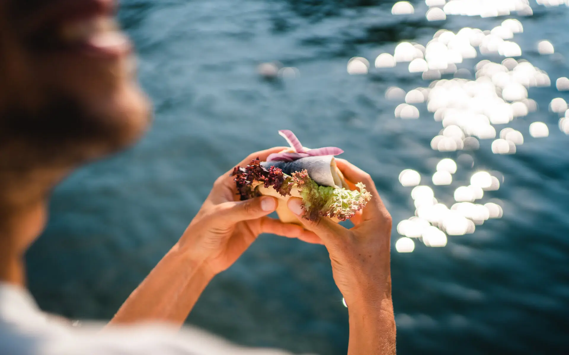 Fischbrötchen Eine Frau steht am Wasser und hält ein Fischbrötchen in ihren Händen. Die Sonne glitzert auf der Wasseroberfläche im Hintergrund.