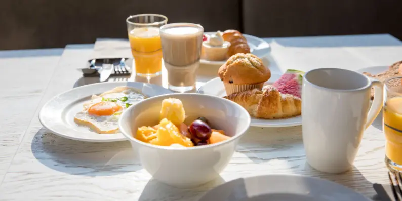 Auf einem Tisch stehen Teller mit verschiedenen Frühstücksspeisen wie Croissants, Früchte, Müsli und Eier sowie Kaffee. Auf einem Tisch stehen Teller mit verschiedenen Frühstücksspeisen wie Croissants, Früchte, Müsli und Eier sowie Kaffee.