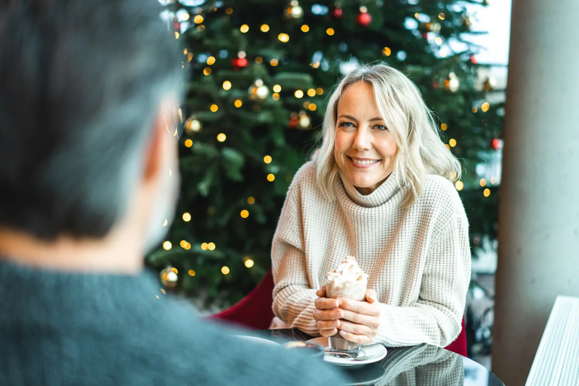 Kuchenstunde Eine lächelnde Frau in einem gemütlichen Pullover sitzt an einem Tisch vor einem festlich geschmückten Weihnachtsbaum. Sie hält ein Getränk mit Sahne in den Händen.