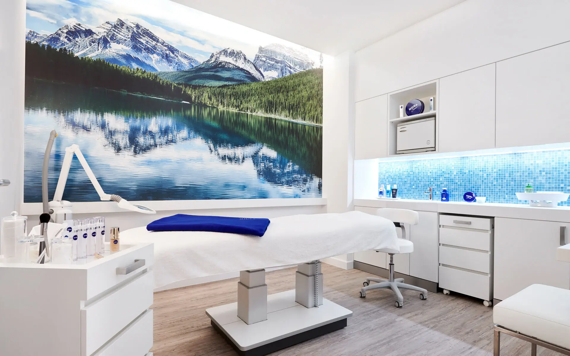 Anwendungsraum im NIVEA Haus Ruhpolding In einem Raum mit hellem Holzboden und weißen Wänden steht vor einem großen Wandbild mit Bergsee-Motiv eine weiße Massageliege auf der ein dunkelblaues gefaltetes Handtuch liegt. Am vorderen linken Rand steht eine weiße Kommode auf der verschiedene Anwendungsprodukte platziert sind.