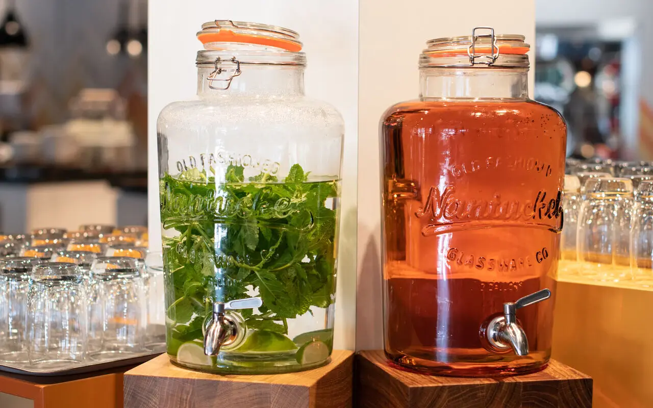 Saftstation Zwei große Glasbehälter mit frischem Wasser und Kräutern stehen nebeneinander auf Holzständern. Der linke Spender enthält klares Wasser mit Minze, während der rechte Spender mit einer rötlichen Flüssigkeit gefüllt ist.Neben den Wasserspendern befinden sich Schalen und Gläser, die für Gäste zur Selbstbedienung.