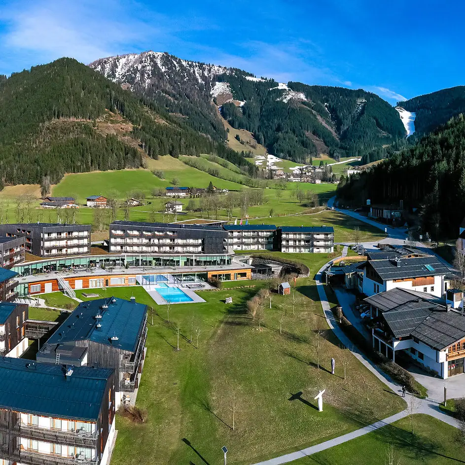 Eine Gruppe von Gebäuden sowie das Hotel mit Pool in einem Tal, umgeben von Bergen im Hintergrund.