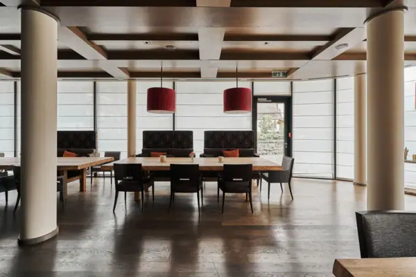 Alpenzeit Ein großer Raum mit Holzboden, großen Tischen und roten Lampen. Die Fenster rund um den räum sind mit Sichtschutz verdeckt.