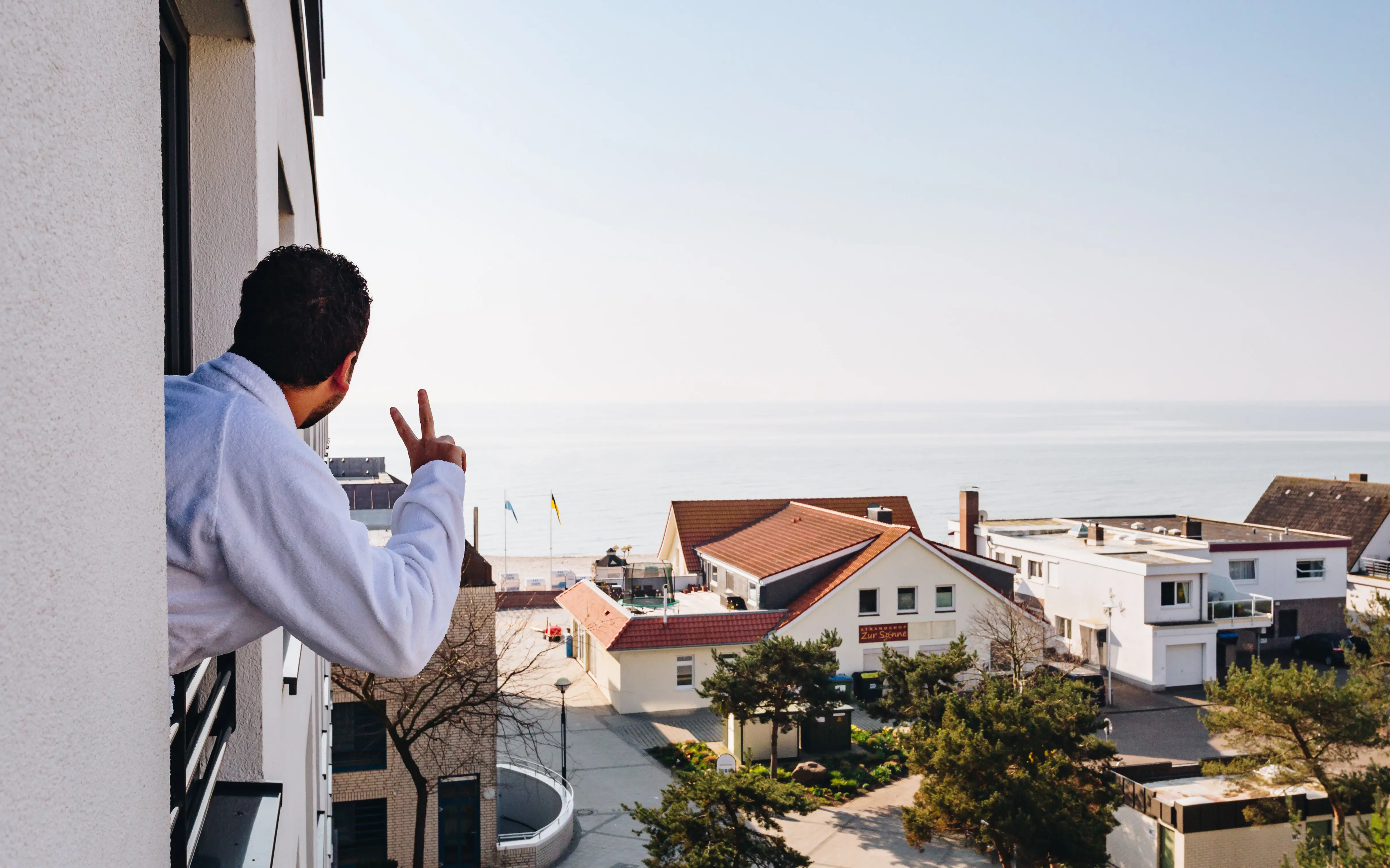 Ein Mann in weißem Bademantel lehnt sich aus einem Fenster und schaut in Richtung Strand, an den sich mehrere Gebäude reihen.