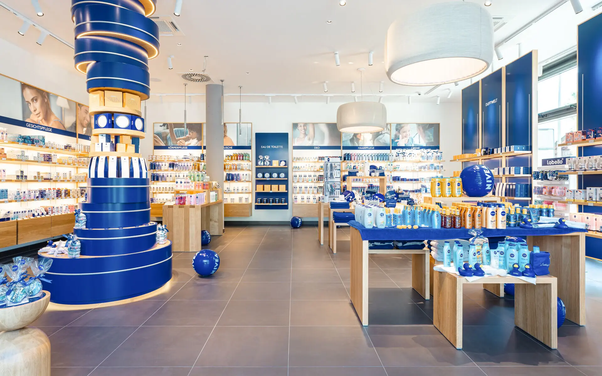 Verkaufsraum des NIVEA Shops Garmisch-Partenkirchen In einem Verkaufsraum mit anthrazitfarbenen Fließen und weißen und dunkelblauen Wänden sind diverse NIVEA Produkte in den Regalen und verschiedenen Verkaufsaufstellern zu erkennen.