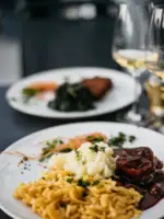 Zwei Teller mit Spätzle, Gemüse und Fleisch in brauner Soße stehen neben zwei Weißweingläsern.