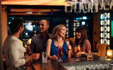 Eine Gruppe von vier Personen unterhält sich gut gelaunt an einer stilvollen Bar. Sie lachen, genießen Cocktails und Longdrinks, während die warme Beleuchtung und das moderne Ambiente eine entspannte und gesellige Atmosphäre schaffen.