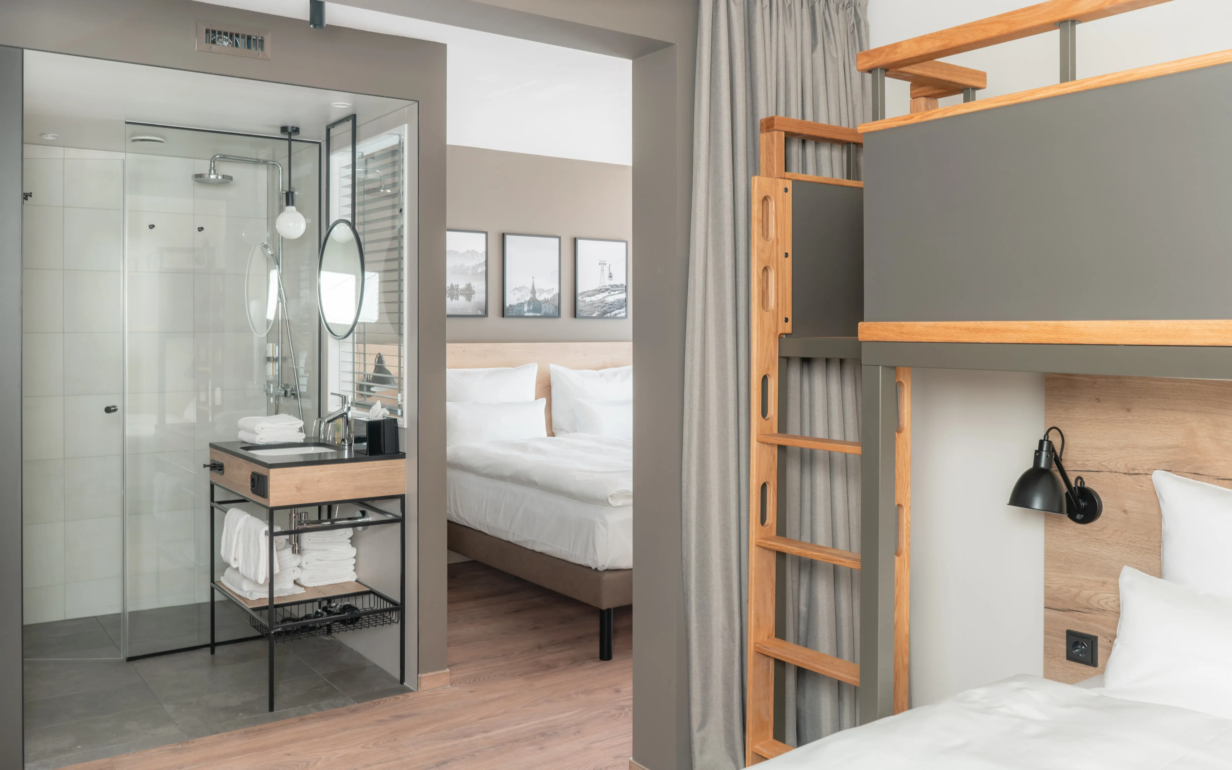 Familienfreundliches Hotelzimmer mit offenem Badezimmerbereich, Doppelbett und modernen Etagenbetten. Das Badezimmer verfügt über eine Glasdusche und ein stilvolles Waschbecken mit Holz- und Metallakzenten.