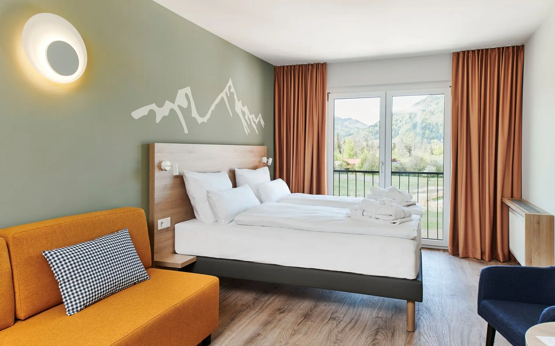 Ein helles Hotelzimmer mit Holzboden in dem ein senffarbenes Sofa uns dahinter ein gemütliches Bett vor eine hellgrünen Wand mit aufgezeichneter Berggruppe stehen. Gegenüber ist ein großes bild von einem See bei Sonnenaufgang an der wand und daneben steht ein blauer Sessel.