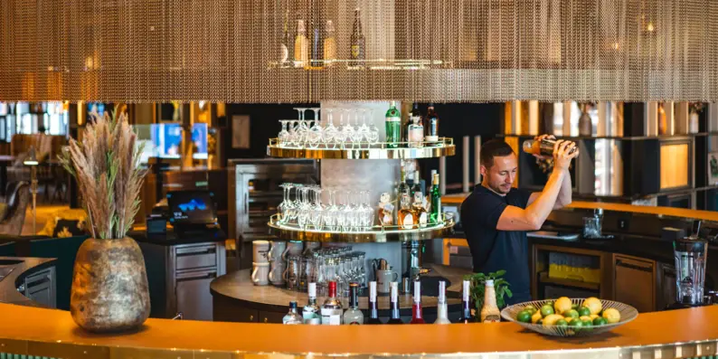 Stilvolle Hotelbar mit einem Barkeeper, der einen Cocktail mixt. Die Bar ist elegant gestaltet, mit goldenen und grünen Akzenten, Gläserregalen und einer großen Vase mit Trockenblumen.Frische Zutaten wie Limetten liegen dekorativ auf der Theke, was eine einladende Atmosphäre schafft. Stilvolle Hotelbar mit einem Barkeeper, der einen Cocktail mixt. Die Bar ist elegant gestaltet, mit goldenen und grünen Akzenten, Gläserregalen und einer großen Vase mit Trockenblumen.Frische Zutaten wie Limetten liegen dekorativ auf der Theke, was eine einladende Atmosphäre schafft.