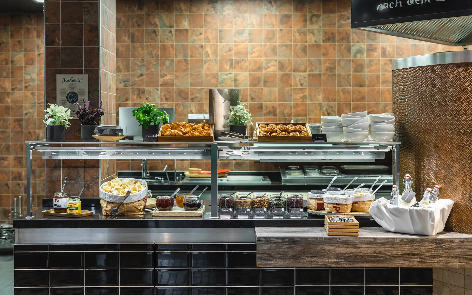 Frühstücksbuffet im aja Bad Saarow Das Frühstücksbuffet im aja Bad Saarow mit Croissants und Brötchen, frischer Marmelade und Müsli. Der Bereich um das Buffet ist in natürlichen Braun- und Grautönen gefliest.