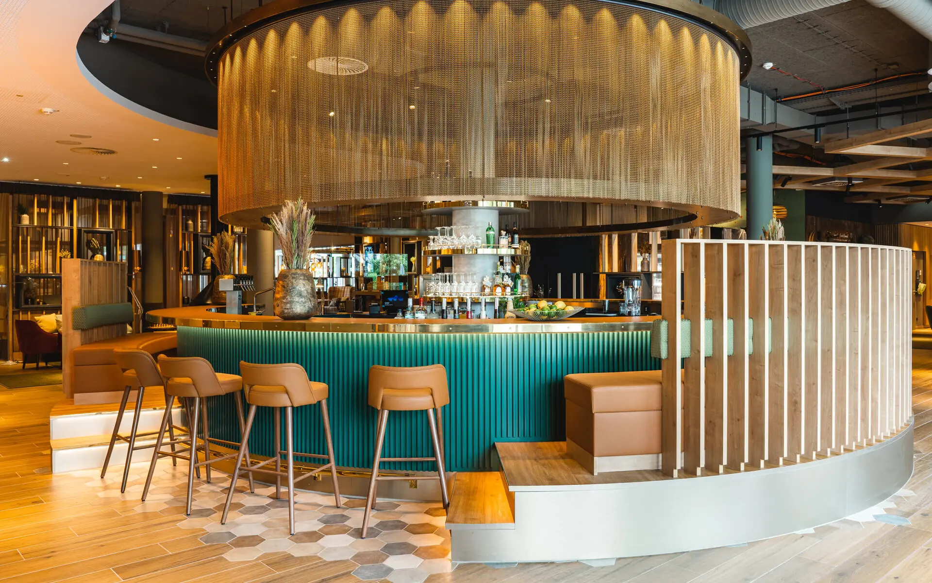 Moderne Hotelbar mit runder Form, goldener Wand und eleganten Holz- sowie grünen Akzenten. Stilvolle Sitzmöglichkeiten und geometrischer Bodenbelag.