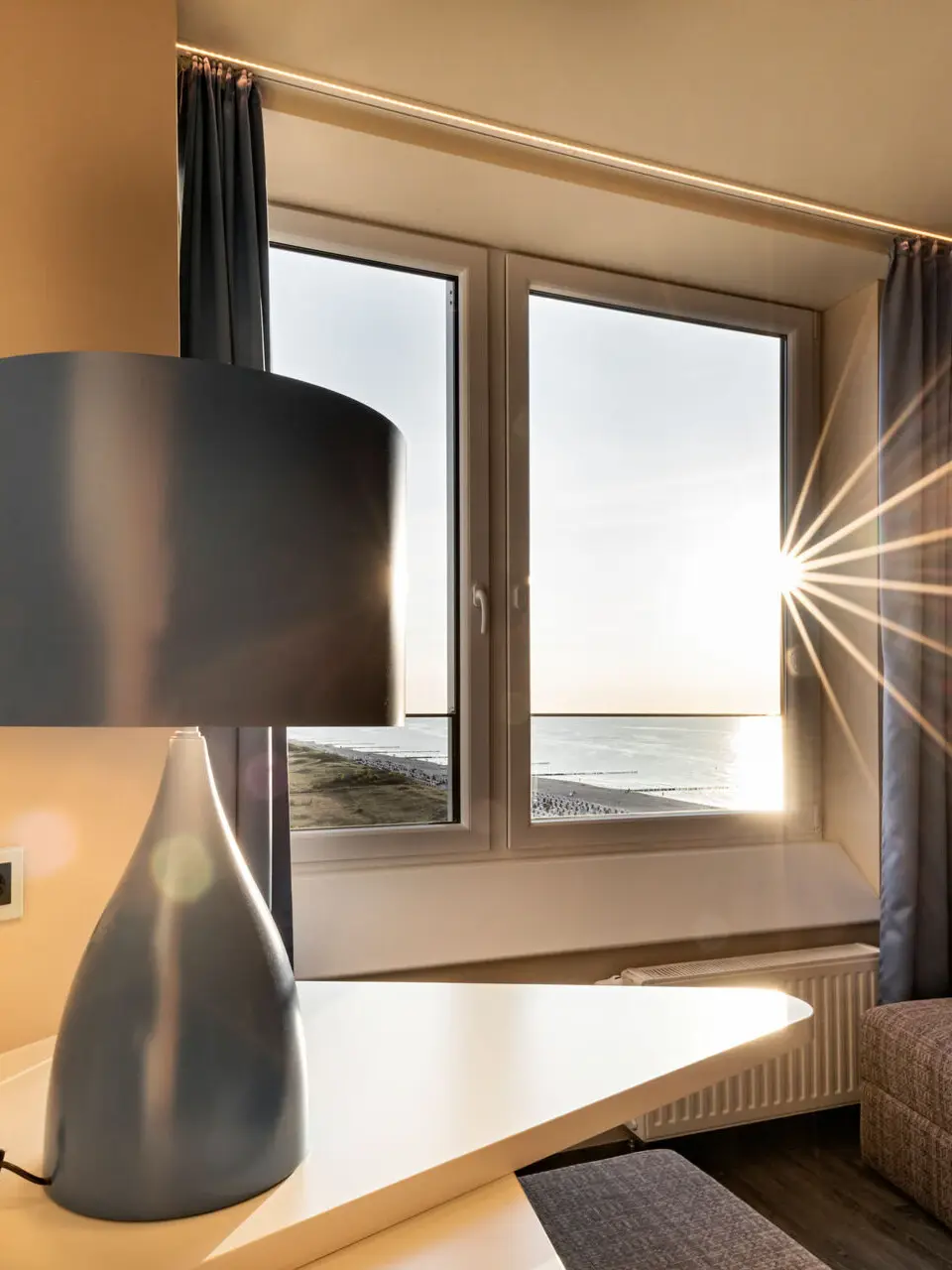 Ein Zimmerausschnitt mit einer graublauen Lampe auf einem Tisch und einem Sofa an einem Fenster, durch das warme Sonnenstrahlen herein scheinen.