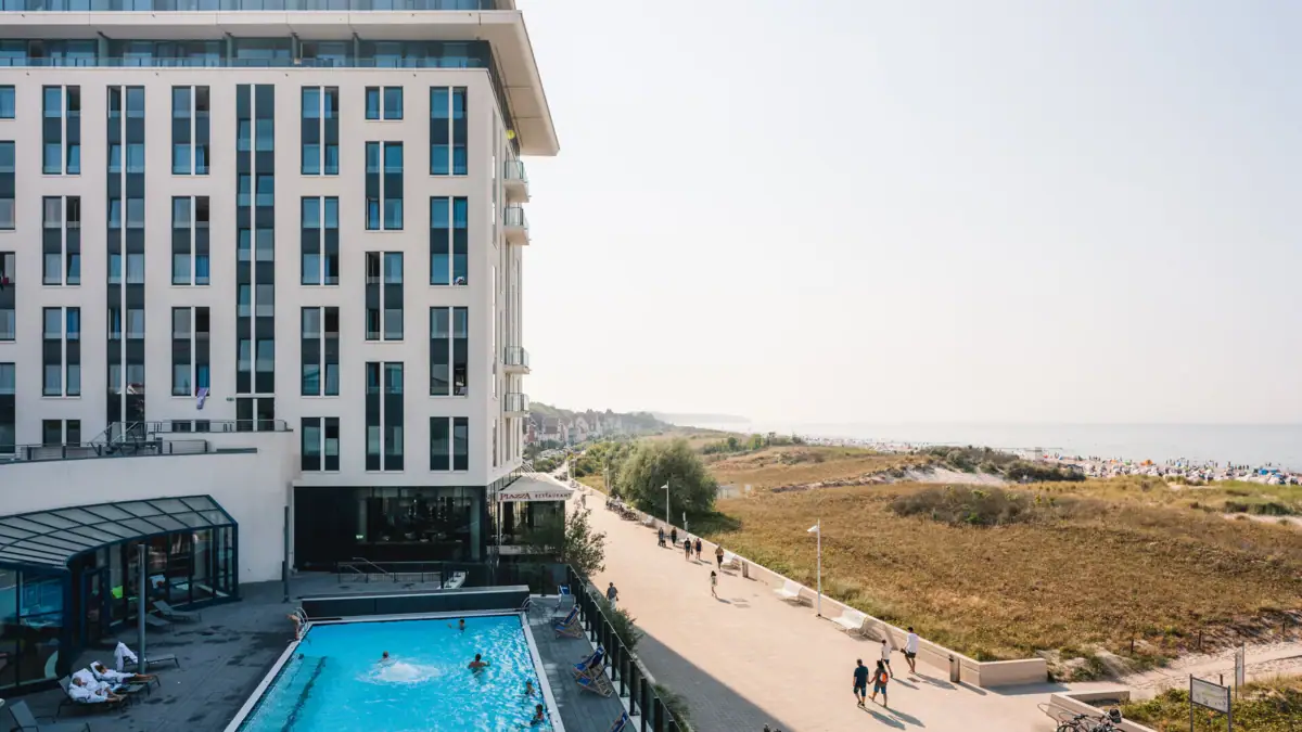 Wellnesshotel aja Warnemünde Ein Gebäude mit einem Pool, an dem Menschen vorbeigehen. Im Hintergrund sind das Meer und der Strand zu sehen.