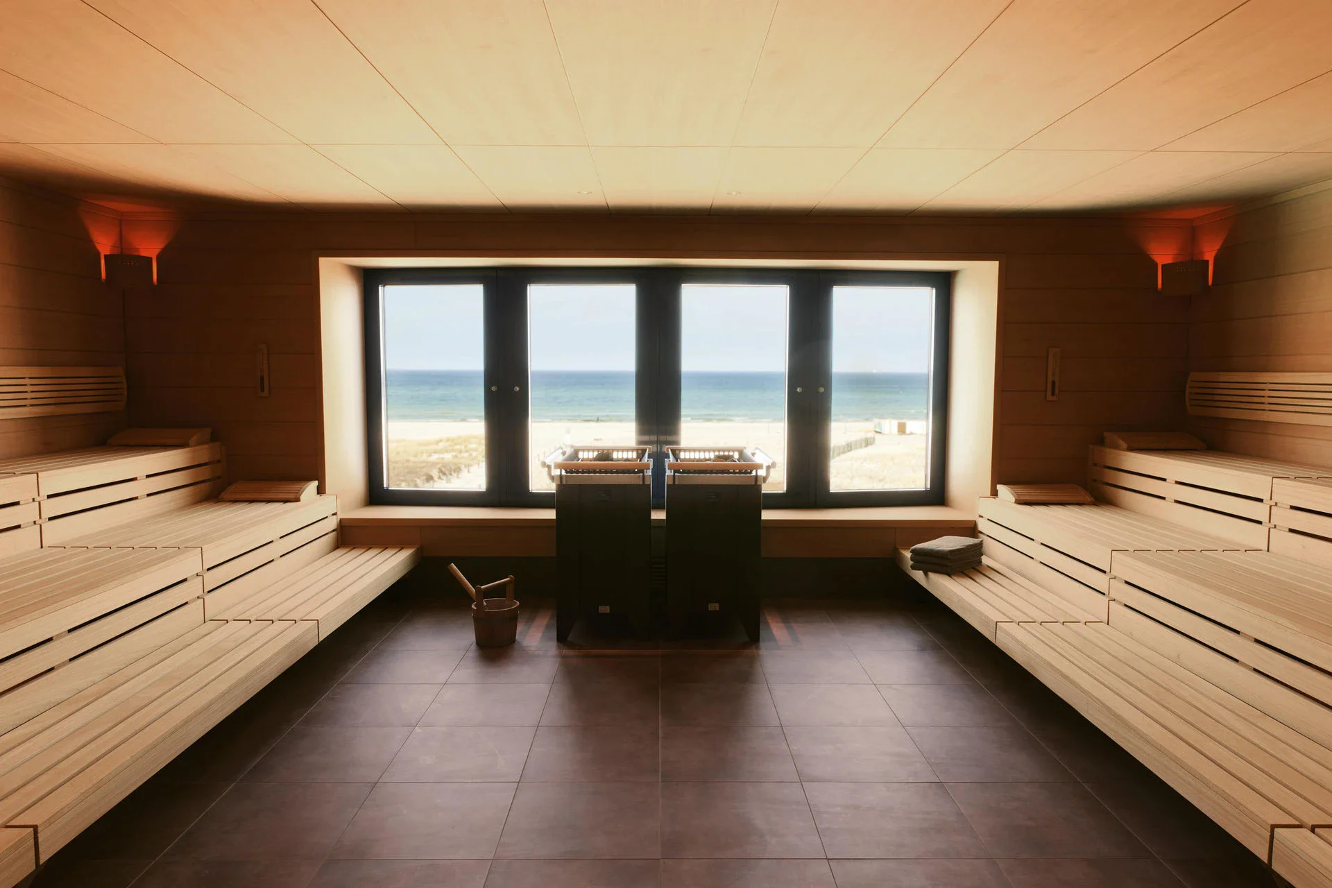 Eine geräumige Holzsauna mit einem großen Fenster mit Blick auf den Strand und das Meer in der Mitte, vor dem zwei schwarze Saunaöfen stehen. Neben dem Fenster, das den Raum mit Tageslicht füllt, befinden sich auf beiden Seiten mehrere Reihen Saunabänke.