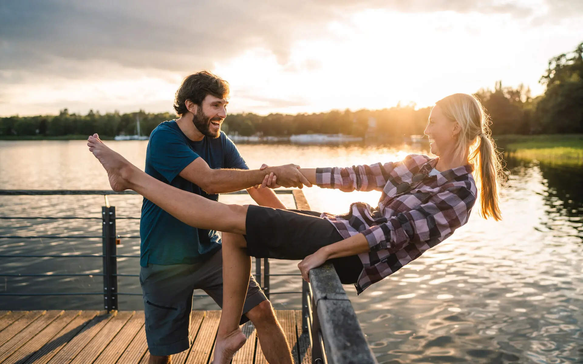 Scharmützelsee Ein lachendes Paar steht auf einem Steg am Scharmützelsee bei Sonnenuntergang. Der Mann hält die Frau spielerischan den Händen fest, während sie sich über das Geländer lehnt.Im Hintergrund spiegelt sich die Sonne im Wasser, und Bäume rahmen die Szenerie ein.