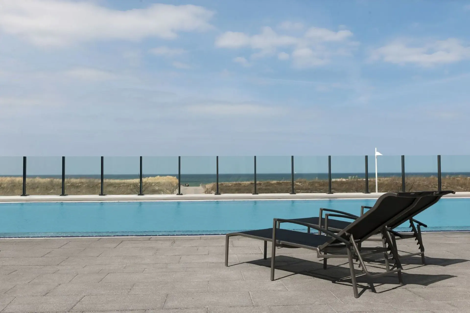 Ein ruhiger Außenpool mit Blick auf die Dünen, den Strand und das Meer. Im Vordergrund stehen zwei Liegen bereit, um die Sonne zu genieße.