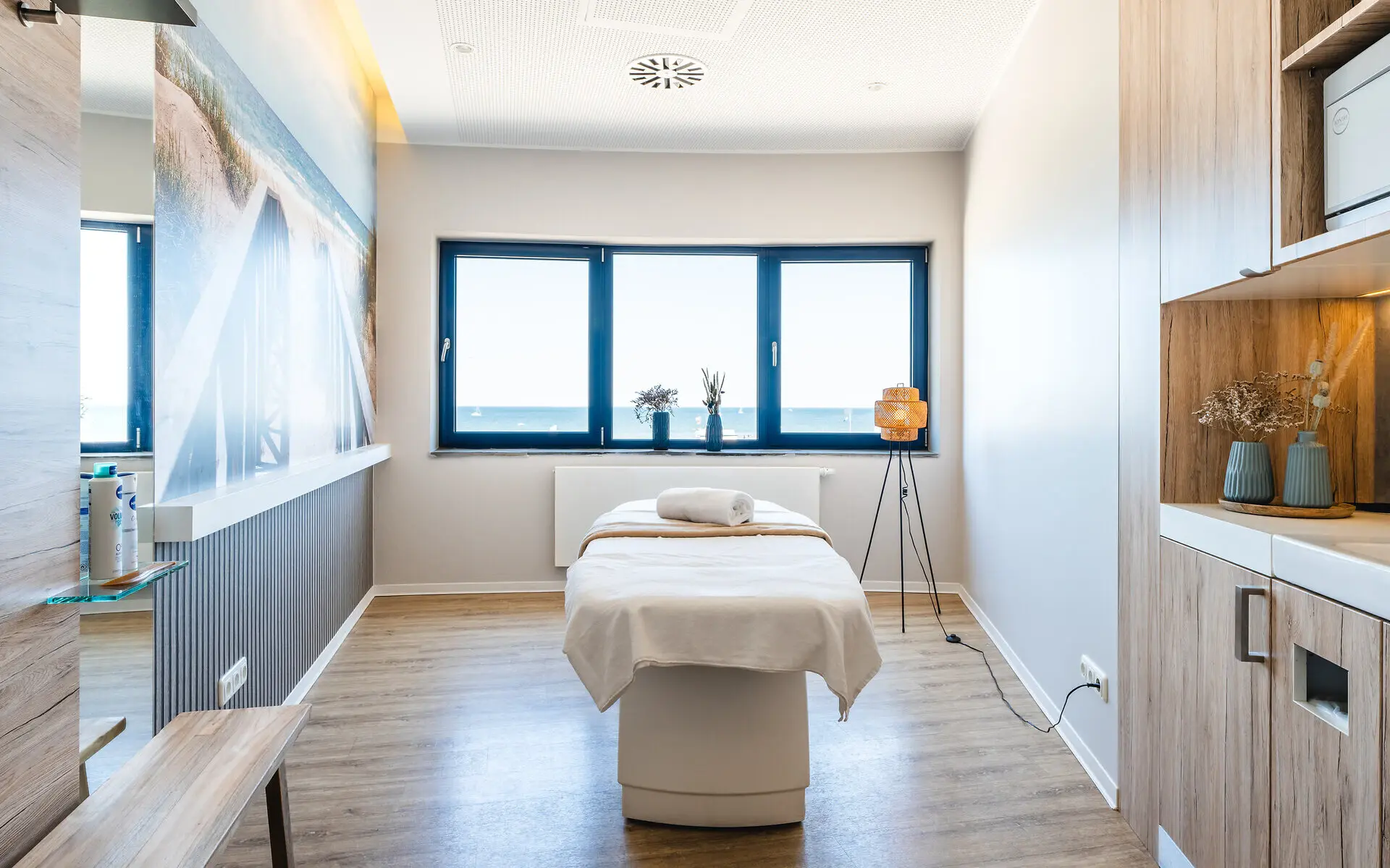 SPA-Anwendungsraum Ein SPA-Anwendungsraum mit Behandlungsliege in der Mitte, einem großen Fenster und Ostseebild an der linken Wand.