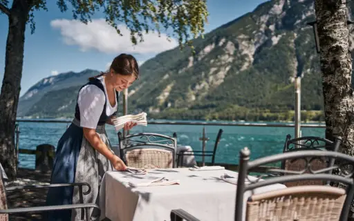 Eine Frau in einem Dirndl deckt einen Tisch an einem See ein. Eine Frau in einem Dirndl deckt einen Tisch an einem See ein.