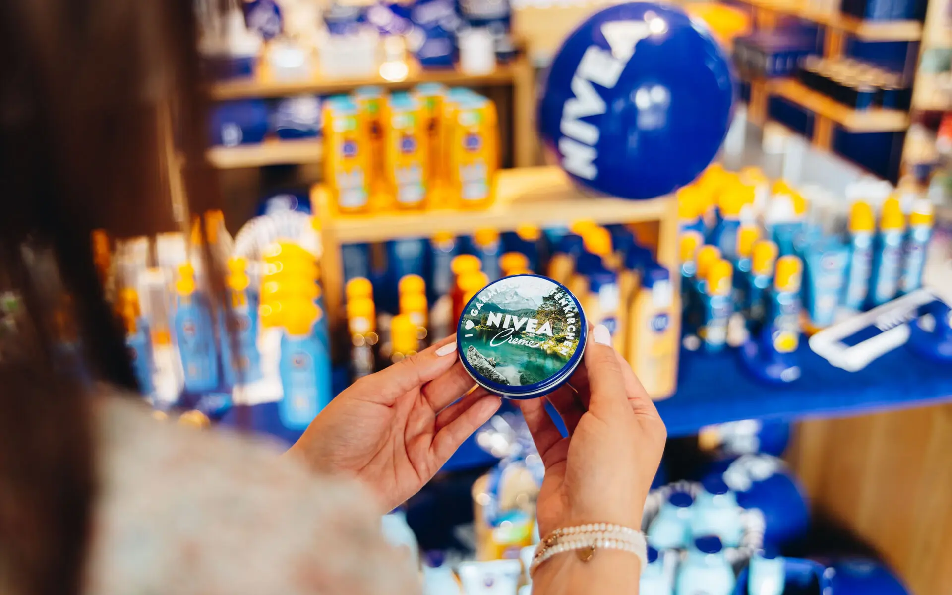 NIVEA Shop Garmisch-Partenkirchen Blick über die Schulter einer dunkelhaarigen Frau, die eine personalisierte Dose einer NIVEA-Creme bedruckt mit einem Bergsee-Motiv in den Händen hält. Im Hintergrund sind verschwommen verschiedene NIVEA-Produkte aufgereiht zu erkennen.