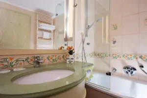 Juniordoppelzimmer landseitig mit Balkon Badezimmer mit Waschbecken und Badewanne, Fliesen an den Wänden, Wasserhahn und Spiegel.