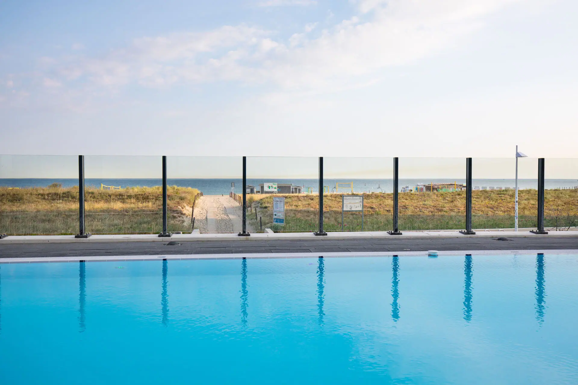 Außenpool im aja Warnemünde Ein Pool mit Zaun und Rasen im Vordergrund, dahinter ein Strand.