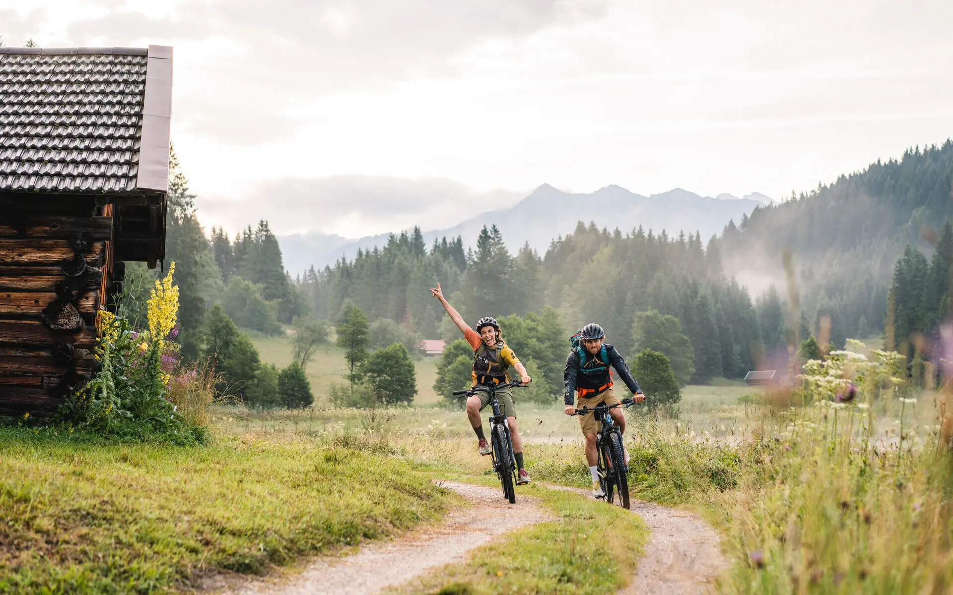 E-Bikes Garmisch-Partenkirchen Zwei Personen fahren auf E-Bikes an einer kleinen Hütte vorbei, im Hintergrund ist eine Waldlandschaft
