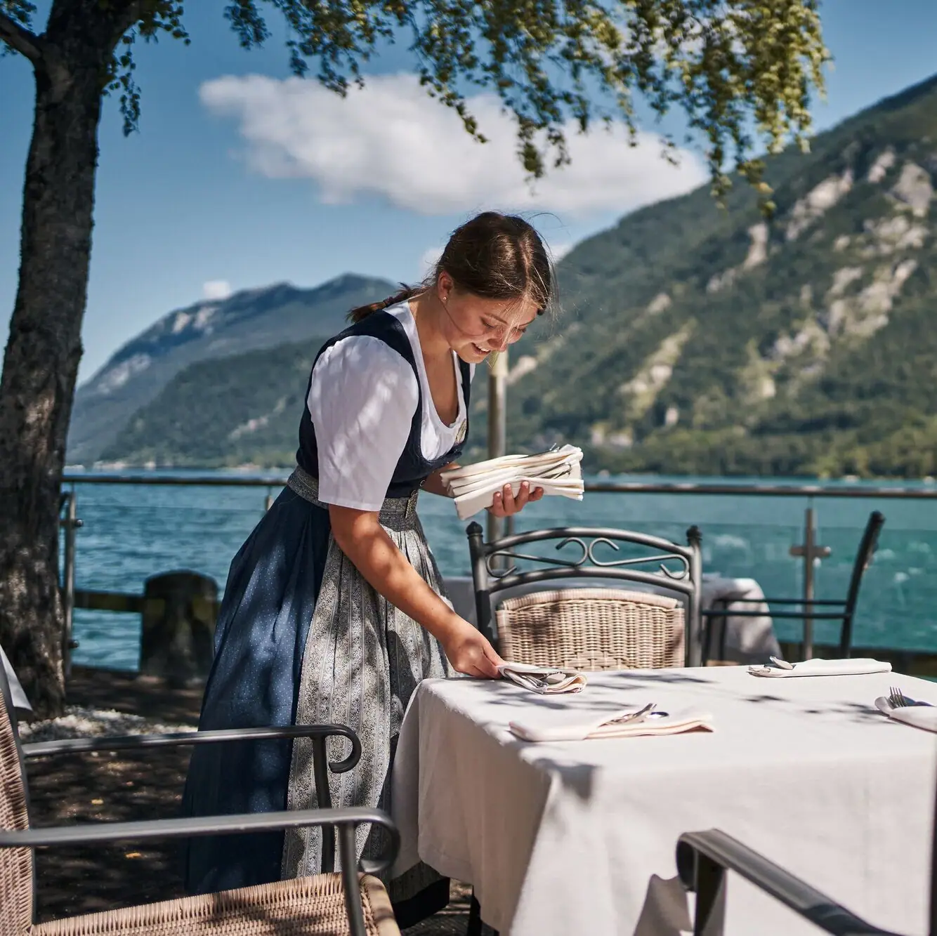 Terrasse am See Eine Frau in einem Kleid an einem Tisch mit einem See im Hintergrund.