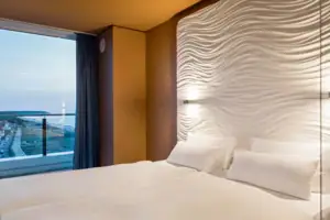Suite im aja Warnemünde Bett mit weißen Laken vor einer Wand mit Fenster mit Blick auf das Meer