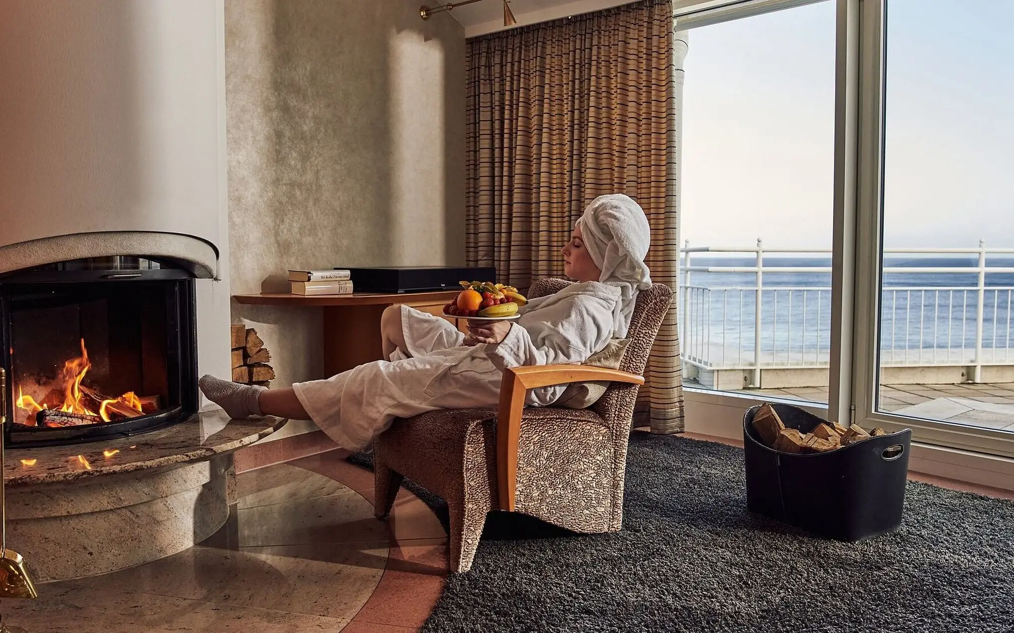 Penthouse-Suite aja Bansin Person in einem Bademantel sitzt auf einem Stuhl neben einem Kamin und einer Schale mit Obst.