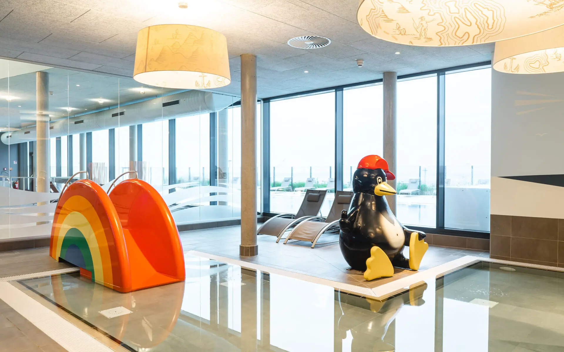 Kinderschwimmbecken aja Travemünde Am Rand eines L-förmigen Kinderschwimmbeckens mit niedriger Wassertiefe steht eine kleine Rutsche in Regenbogenform und eine sitzende Pinguin-Figur mit einer roten Mütze auf dem Kopf. Im Hintergrund stehen zwei nicht besetzte Poolliegen vor einem großen Panoramafenster mit Blick auf den Außenbereich.