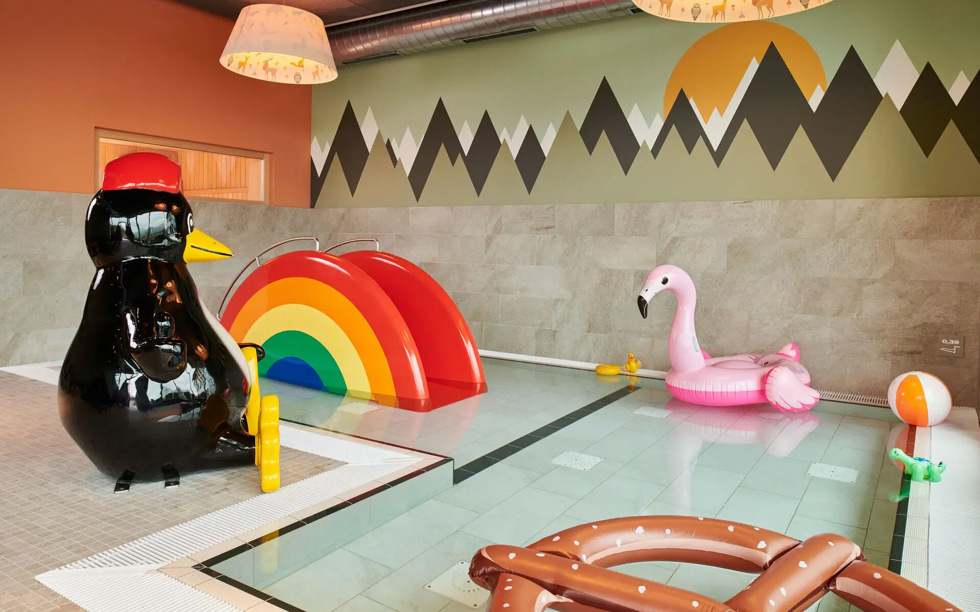 Kinderschwimmbecken aja Ruhpolding Am Rand eines L-förmigen Kinderschwimmbeckens mit niedriger Wassertiefe steht eine sitzende Pinguin-Figur mit einer roten Mütze auf dem Kopf. Im hintern Bereich des Beckens steht eine kleine Rutsche in Regenbogenform und Im vorderen Bereich schwimmt ein aufblasbarer Schwimmreifen in Flamingo-Form sowie ein weiß-orange gestreifter Wasserball. Am vorderen Beckenrand liegt eine große aufblasbare Brezel.