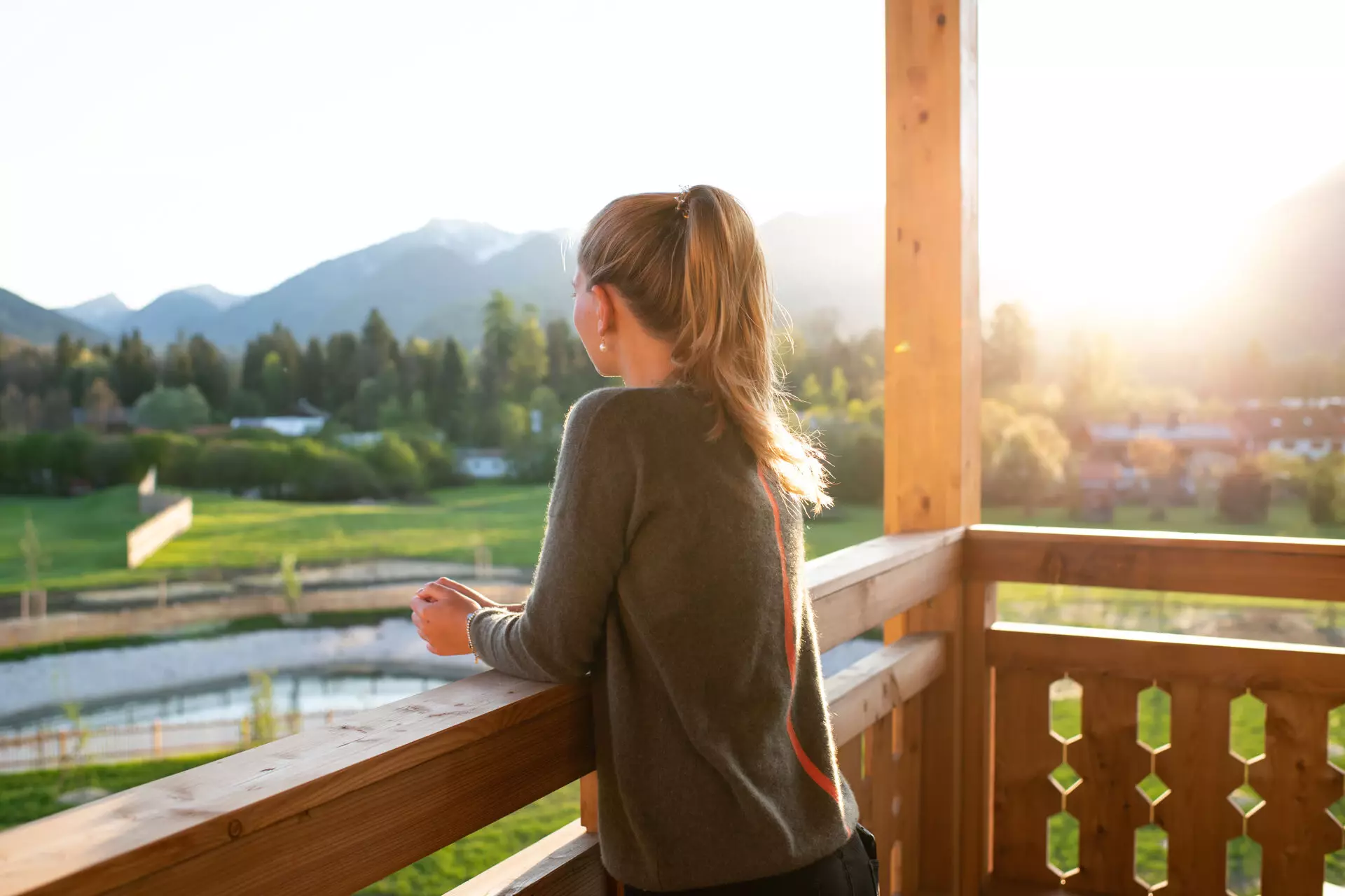 Eine Frau steht auf einem Holzbalkon und genießt den weiten Ausblick auf eine sonnige Berglandschaft. Die warmen Strahlen der aufgehenden Sonne tauchen die Szene in ein weiches Licht.
