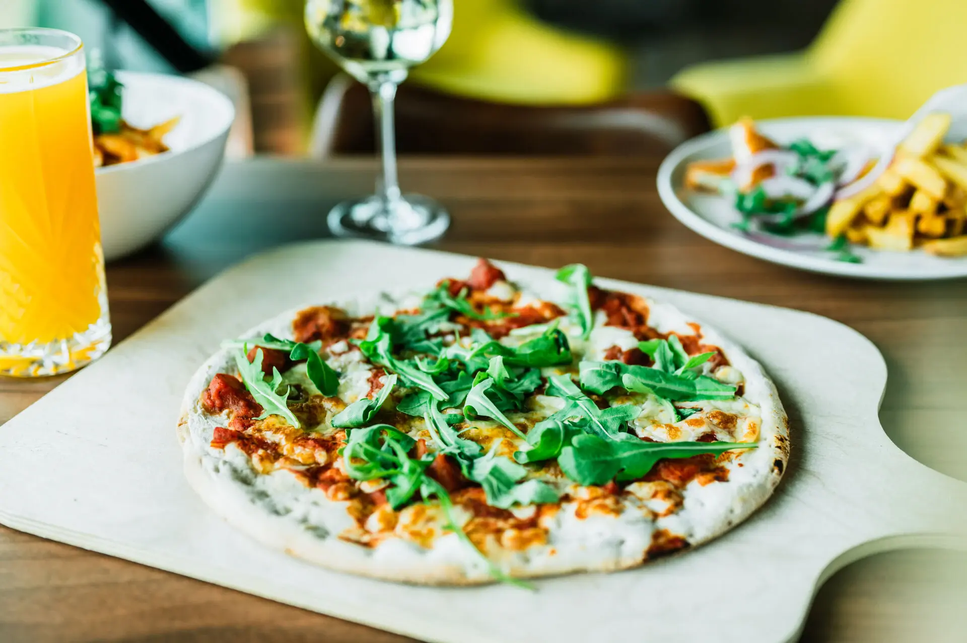 Frisch gebackene Pizza mit Rucola auf einem Holzbrett, begleitet von einem Glas Orangensaft und weiteren Gerichten im Hintergrund, angerichtet in einem modernen Restaurant.
