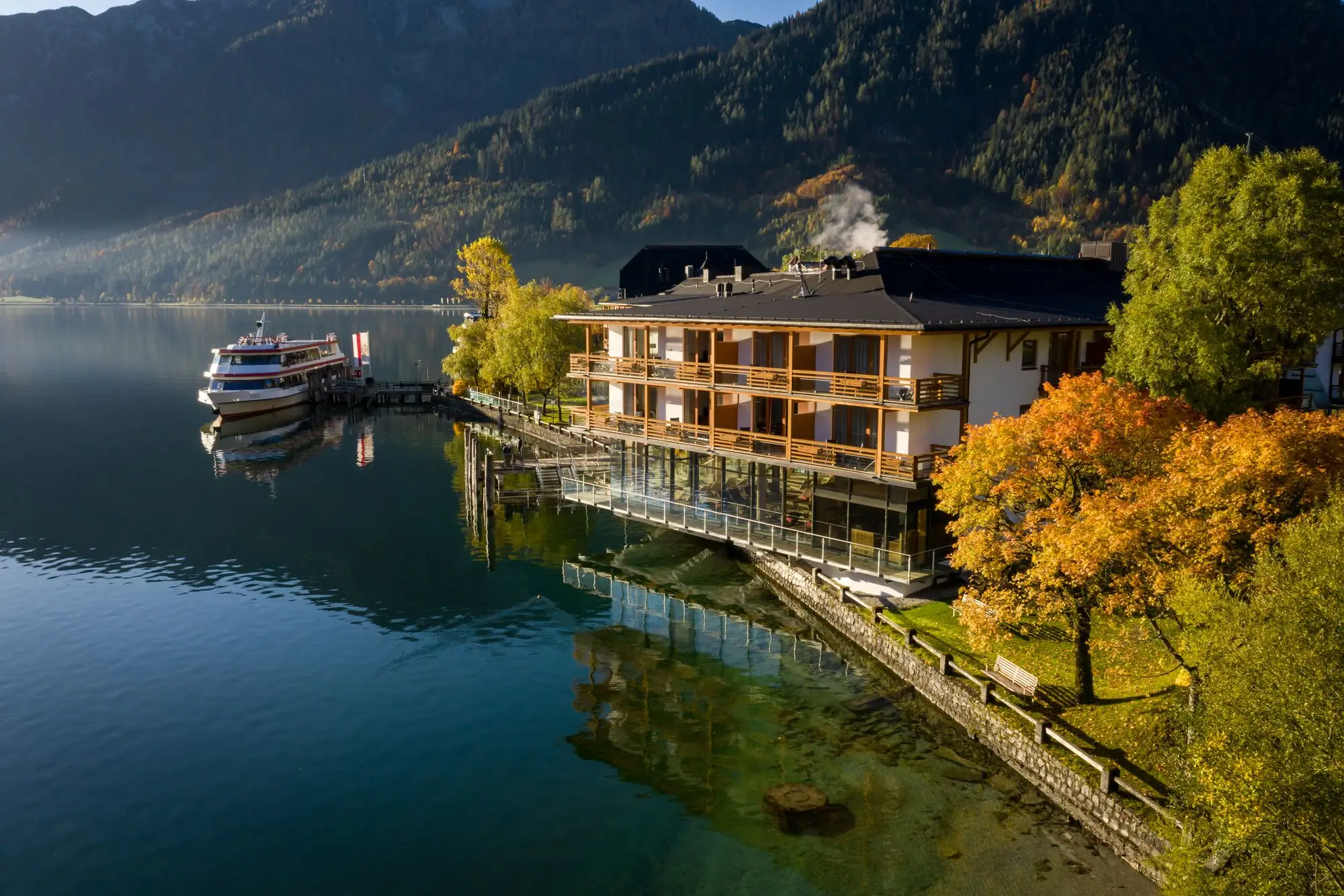 aja Fürstenhaus am Achensee Gebäude am Wasser mit Bäumen und Bergen im Hintergrund.
