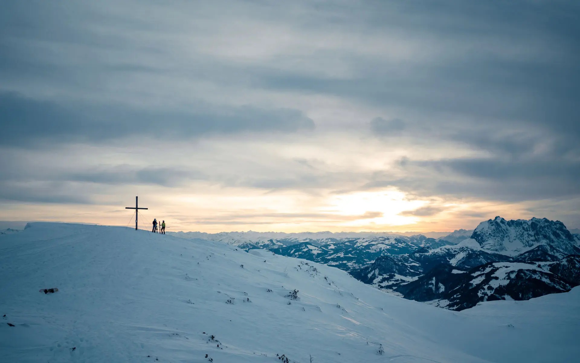 Auf einem weiten schneebedeckten Berg steht in der ferne ein Kreuz, neben dem zwei Personen stehen. Am Horizont geht die Sonne auf und es erstrecken sich weite schneebedeckte Berggruppen.