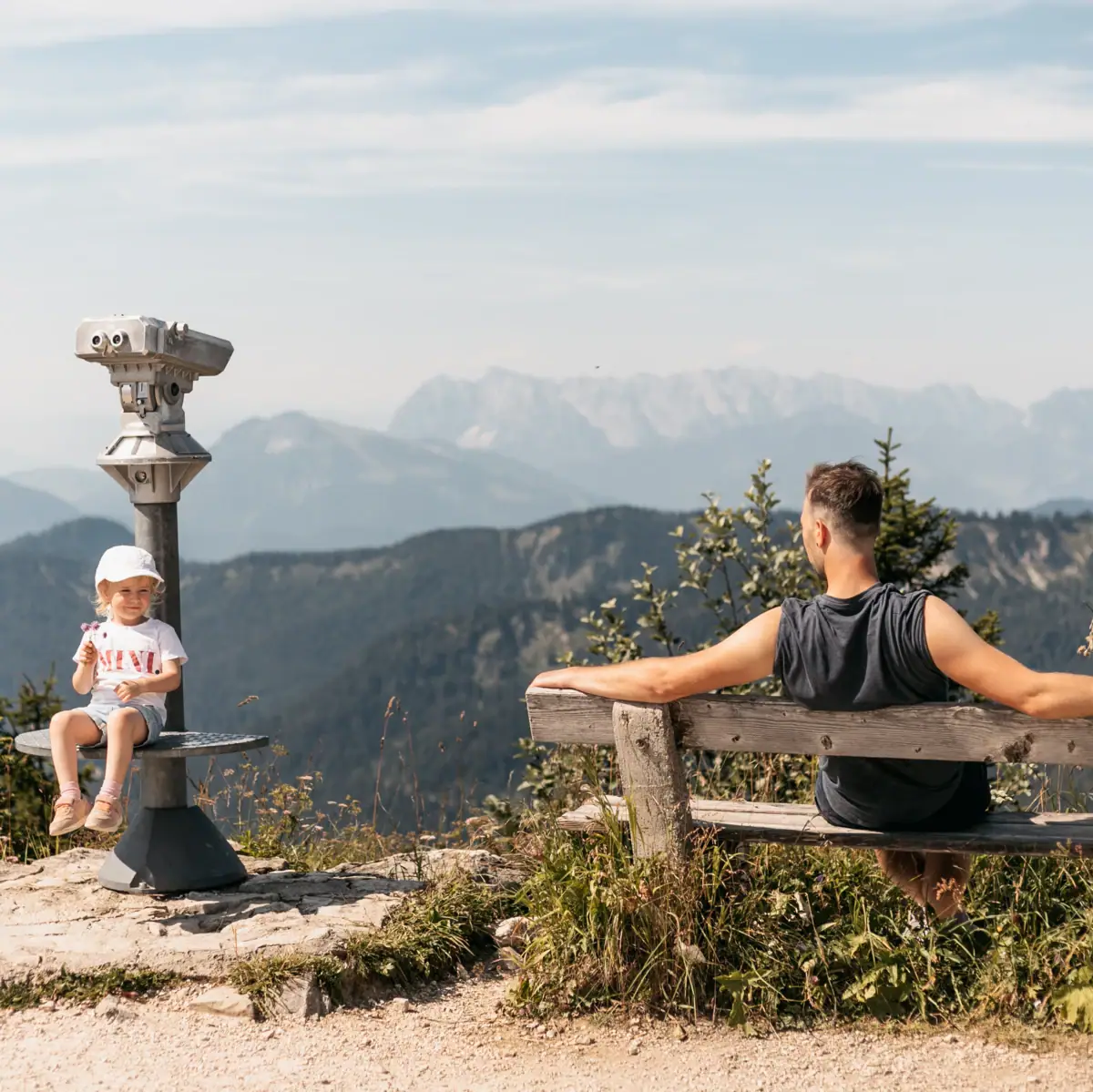 Aussicht vom Berg Ein Mann und ein Kind sitzen auf einer Bank mit Blick auf Berge.