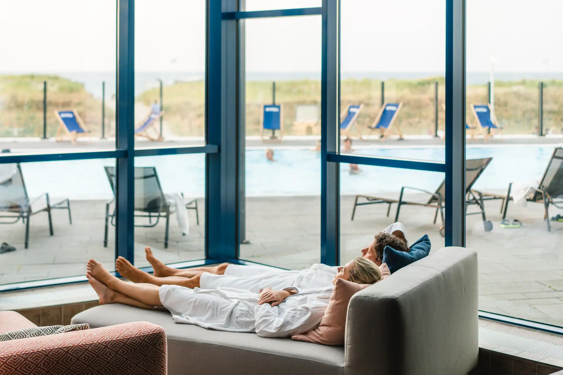 Ruheligen im aja Warnemünde Ein Paar liegt auf einem Wellness-Sofa mit Blick raus auf den Außenpool.