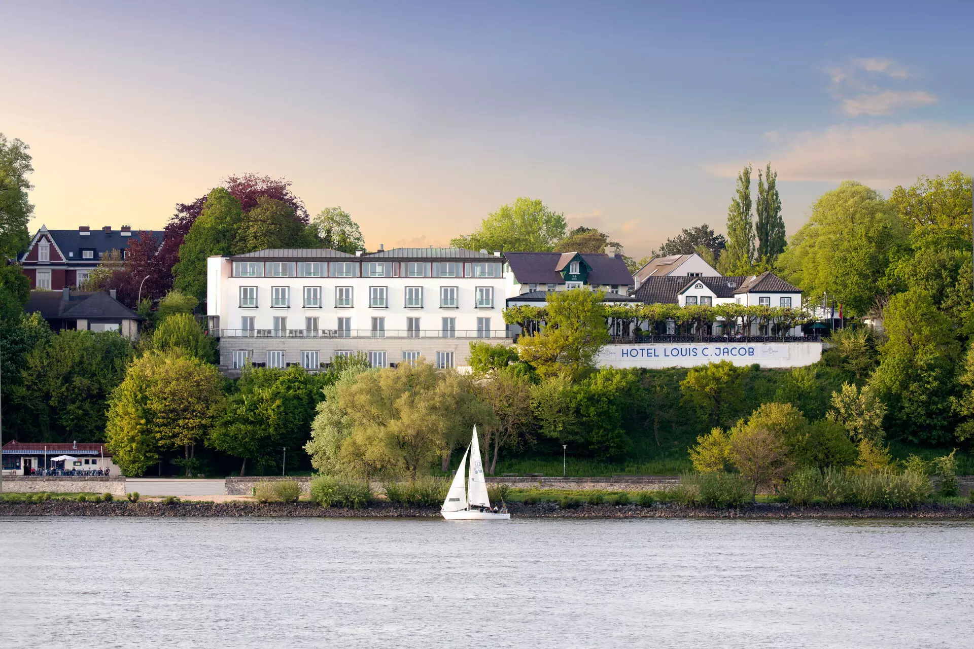 Louis C. Jacob Außenansicht des Hotel Louis C. Jacob, idyllisch gelegen an der Elbe, umgeben von üppigem Grün, mit einer Terrasse und einem Segelboot im Vordergrund, das auf dem Wasser vorbeifährt.