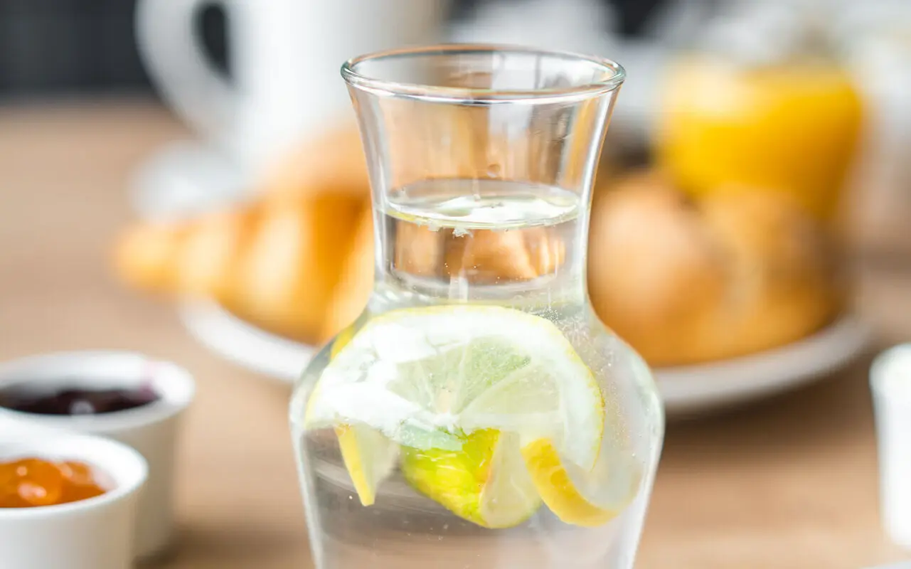 Wasser Eine Wasserkaraffe mit Zitronenscheiben drin. Im Hintergrund steht ein verschwommener Teller mit Brötchen.