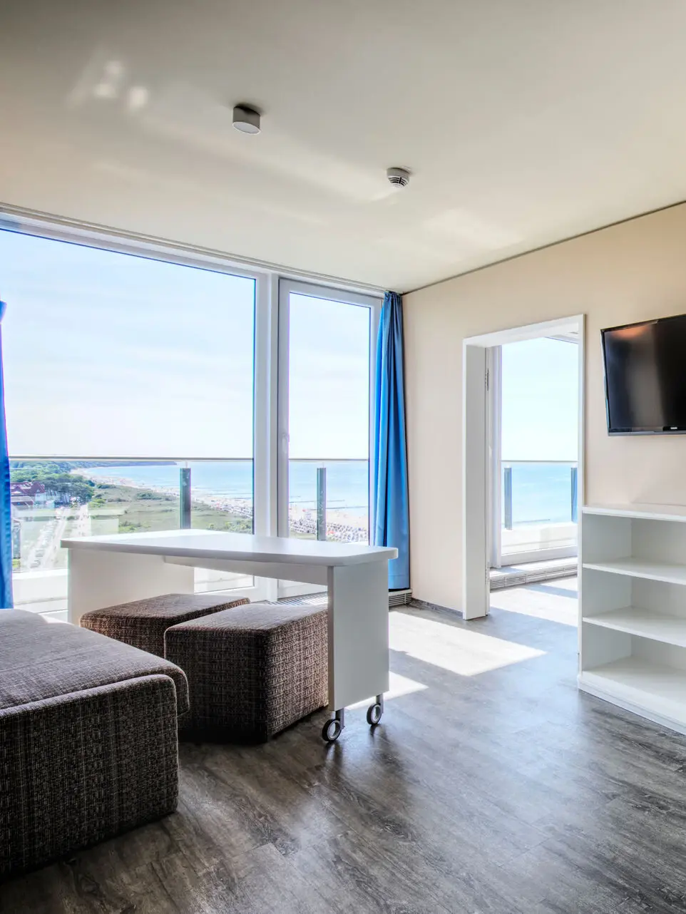 Ein heller Raum mit braunem Sofa und einem weißen Tisch auf Rollen davor, unter em zwei Hocker stehen. Durch große Fenster fällt viel Tageslicht in den Raum und eine Tür führt in einen weiteren Raum.