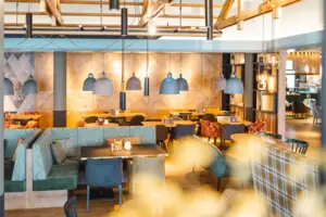 Eleganter Restaurantbereich mit warmen Holztönen und modernen Sitzgelegenheiten. Verschiedene Tische und Sitzbänke sind gemütlich angeordnet, ergänzt durch stilvolle Hängelampen in Blau- und Gelbtönen.