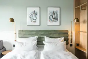 Zimmer im aja Warnemünde Ein Doppelbett mit weißen Kissen und einem grünen Kopfteil, flankiert von zwei Lampen und einem offenen Schrank